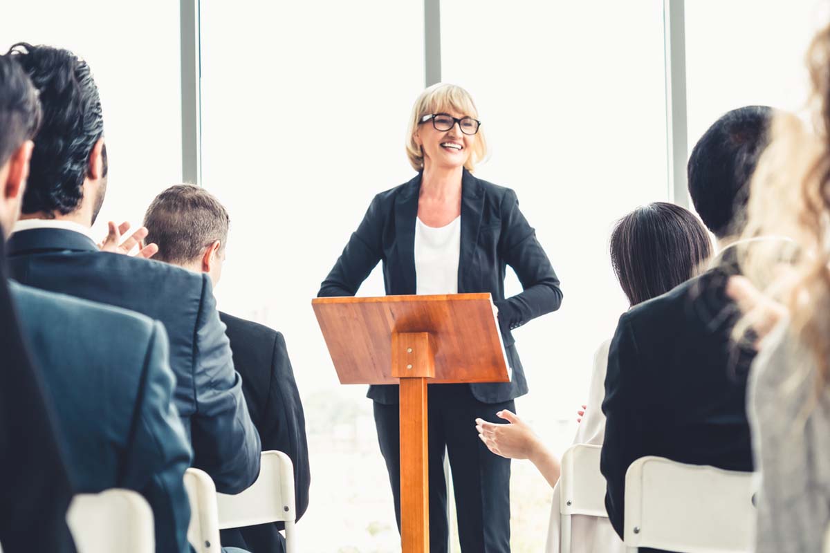 Older woman standing at a podium speaking to an audience at a conference in an effort to boost lead generation